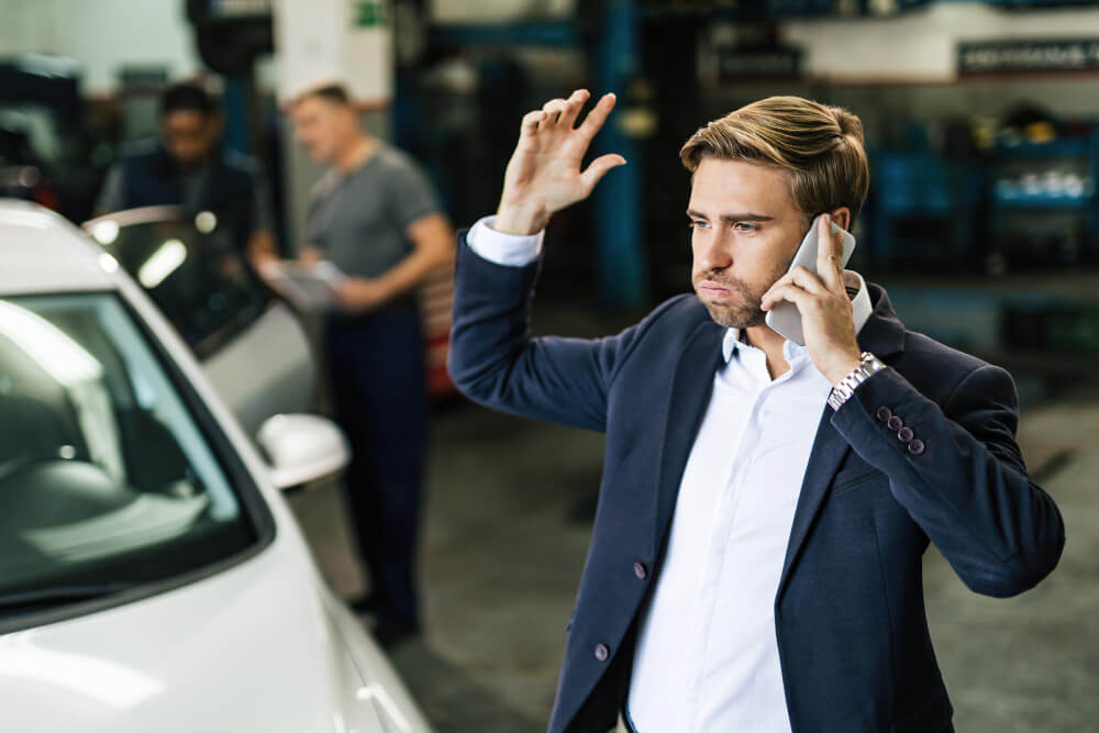 Délai de livraison de votre voiture d'occasion non respecté : quels sont vos droits ? 2 Client l'air impatient, devant un garage automobile avec une voiture d'occasion en arrière-plan.