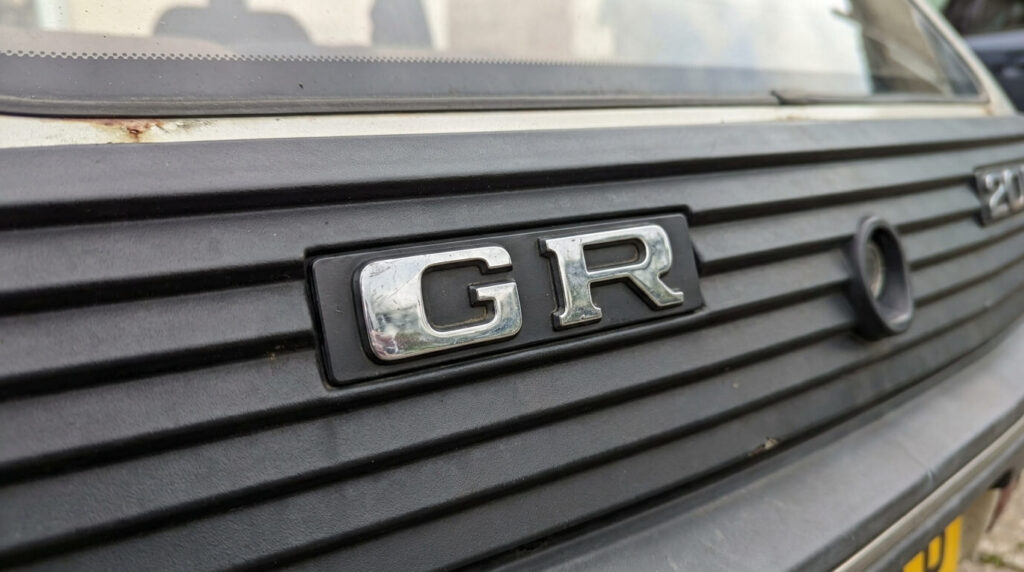 Badges Peugeot 205, 309 et 405 expliqués : GL, GR, SR, GTI, et plus 3 Gros plan sur le badge en plastique chromé GR fixé sur la râpe de hayon arrière d'une Peugeot 205 d'époque.