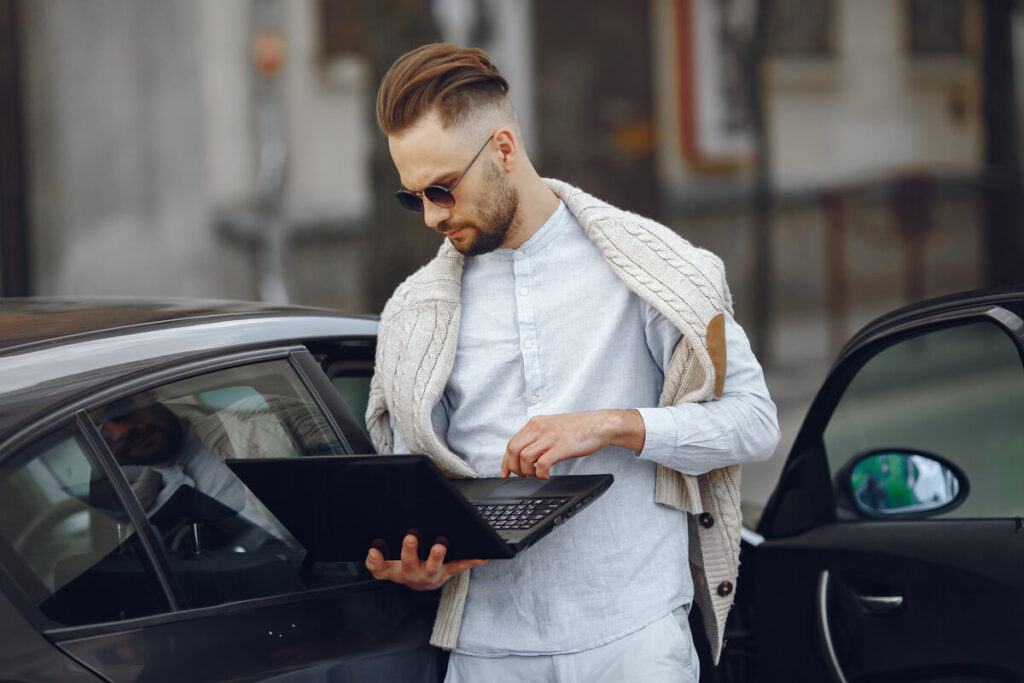 Les cyber-risques des voitures connectées : ce que les conducteurs doivent savoir 3 Un hacker sur un ordinateur portable à côté d'une voiture moderne, symbolisant le risque de piratage à distance.