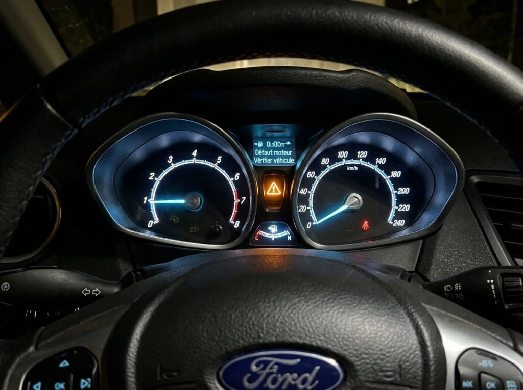 Voyant lumineux d'alerte orange avec point d'exclamation au centre d'un triangle sur le compteur d'une Ford Fiesta.